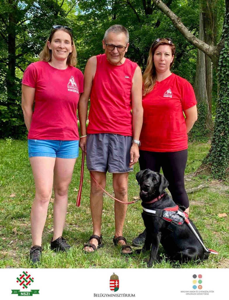 A fotón balról jobbra Végel Gabriella, Nagy János és Bajnóczy Enikő áll. Mindhárman piros pólót
viselnek. Előttük Zorka ül piros MVGYOSZ-hámban, sokatmondó, okos tekintettel néz a kamerába.