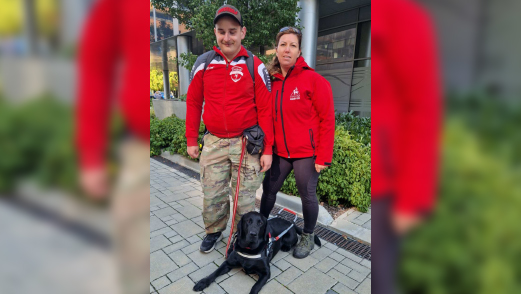 A képen Kovács Attila és Bajnóczy Enikő látható. Mirkó, a fekete labrador, lábuk előtt fekszik piros MVGYOSZ feliratú vakvezetőhámban.