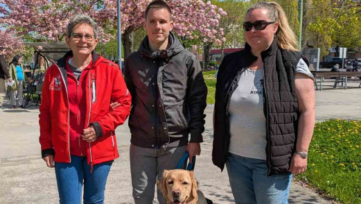 Kiss-Tóth Zoltán Ferencné Marika, Nagy Dávid és Rózsa Kata a tavaszi napsütésben. Dávid lábánál a hosszúszőrű labrador, Elvis ül „mosolyogva”. Háttérben virágzó fák és egy park részlete látható.